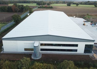 Aerial view of a large, modern warehouse building with a white roof and surrounding greenery.